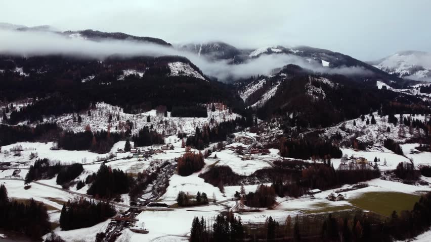 Aerial drone view of snow-covered valley with scattered houses, dense forests, and hills under low clouds covering mountain peaks in Trentino-Alto Adige