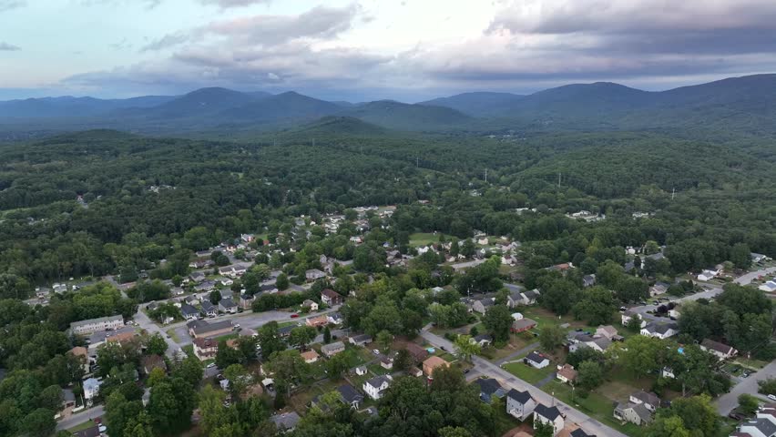 Drone wide shot of Waynesboro, Virginia, with residential neighborhoods surrounded by lush forests. Blue Ridge Mountains rising in background under dramatic cloudy sky. Quiet suburbia district.