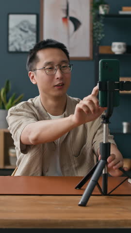 Vertical shot of young male content creator of Asian ethnicity talking on camera of smartphone in tripod while vlogging at home