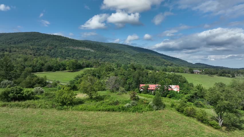 Cars driving on rural road between green farm fields and farmstead. Sunny summer day in Virginia. Blue ridge mountains in background. Aerial rising wide shot.