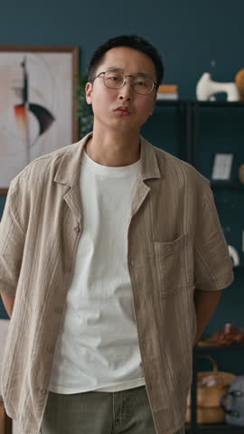 Vertical portrait of young Asian man confidently looking at camera in modern living room with abstract pictures in background