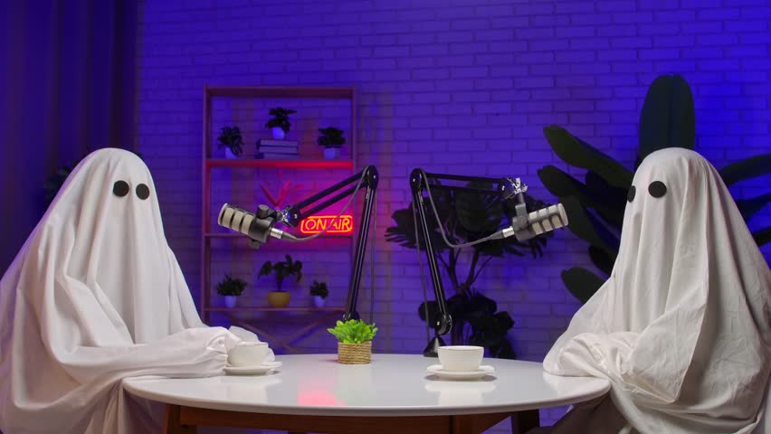 Two people dressed as ghosts, wearing white sheets and headphones, record a podcast at a studio table with microphones. Perfect for Halloween, comedy, and entertainment.