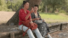 Two women sharing a relaxing lunch moment, sitting under verdant tree shade, enjoying engaging conversation in peaceful outdoor park setting during sunny summer day - Powered by Shutterstock - Get 15% off with code: PIKWIZARD15