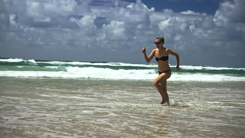A stunning super slow-motion shot captures a slim young woman in a swimsuit running along the shoreline, kicking up water sprays against crashing ocean waves and cloud-dotted skies.