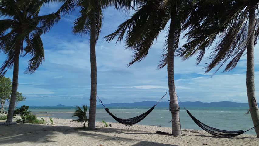 Drone footage capturing hammocks strung between palm trees on a sandy beach in Koh Phangan Thailand with turquoise sea, tropical coastline, and tranquil summer atmosphere.