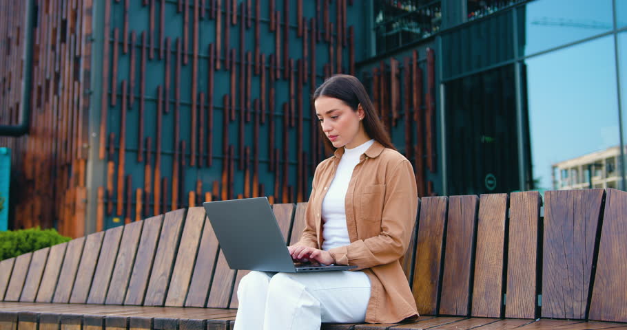 Young business woman remotely on her computer while sitting on bench during lunch break about modern office building, outdoors