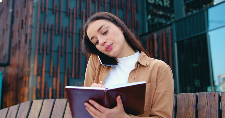 Close up of self employed female making phone call on mobile phone and taking notes while working remotely while sitting on bench about modern office building, outdoors