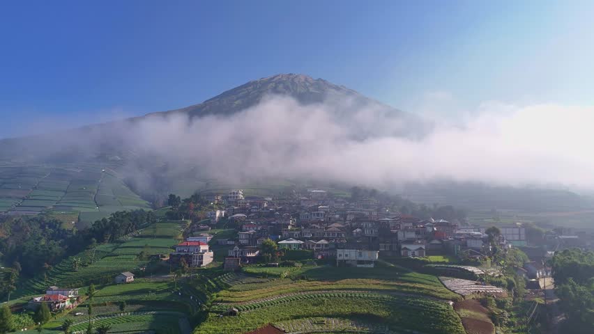 Drone view showing mountain slope with terraced fields, village houses, and a towering mountain covered by clouds.