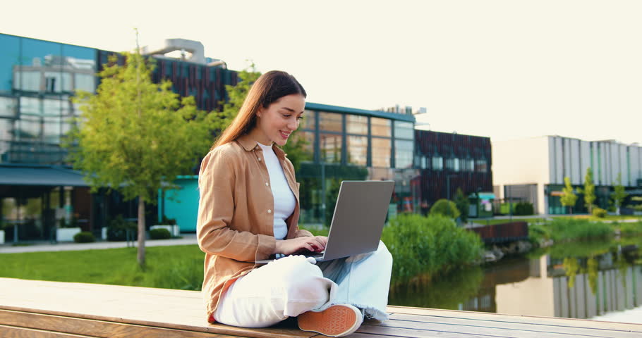 Beautiful and smiling Caucasian girl sitting cross-legged on bench in city park, working remotely on her laptop while enjoying the fresh air and peaceful surroundings