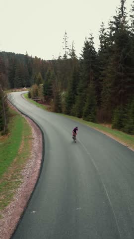 Stylish female cyclist in violet jersey and black helmet riding road bicycle uphill along new empty asphalt road in mountains, aerial view. Vertical video