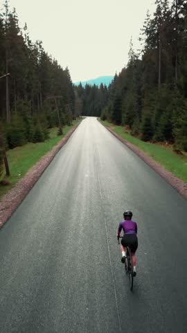 Female cyclist in violet cycling apparel and black helmet rides on road bicycle in national park in mountains. Aerial drone shot of woman cycling beautiful epic landscape. Vertical video