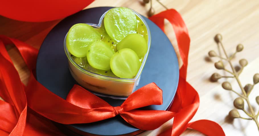 Heart shaped dessert featuring green grapes and kiwi slices, resting on dark plate with red ribbon, expressing romantic valentine's day sweetness