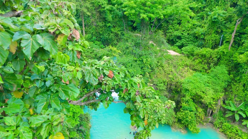 Aerial view of Kawasan Falls, a multi-tiered waterfall cascading into a bright turquoise river surrounded by lush tropical jungle, with people swimming, Cebu, Philippines.