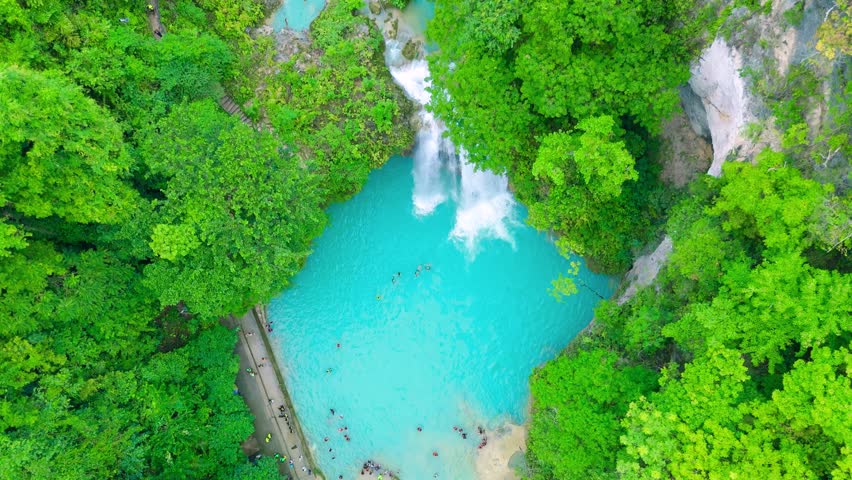 Aerial multi-tiered waterfall cascading into a turquoise pool, surrounded by green jungle foliage with people swimming, Kawasan Falls, Philippines.