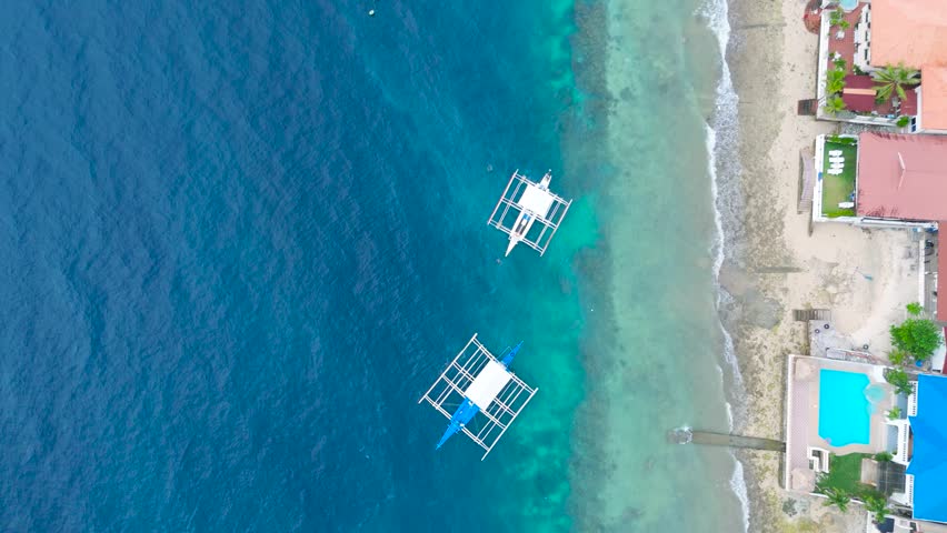 Aerial view of traditional outrigger boats floating on turquoise ocean water near a sandy beach and coastal resorts in Moalboal, Cebu, Philippines.