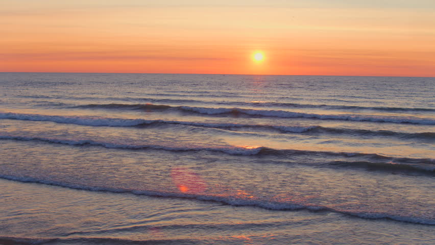 Drone aerial of sunrise over Lake Michigan with gentle waves rolling onto a sandy beach as birds fly past a pier under a glowing horizon on a clear morning.