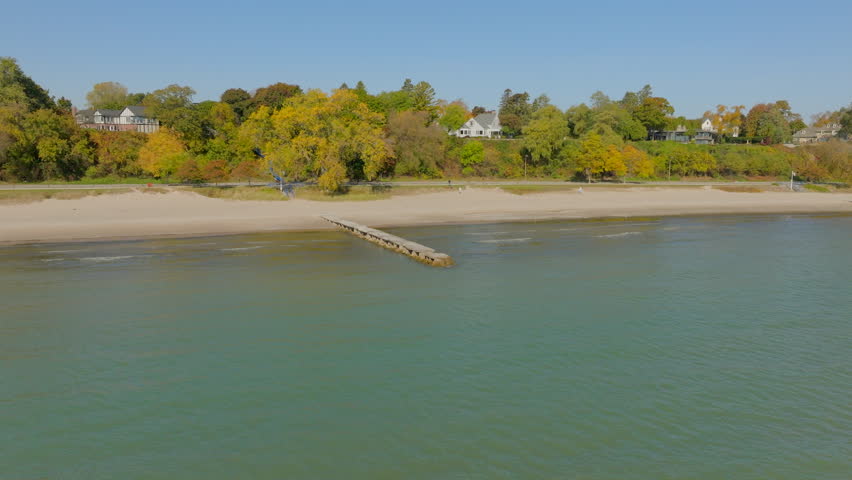 Drone aerial over Lake Michigan toward the shoreline with a pier and a house surrounded by colorful autumn trees under a clear blue sky on a bright day.