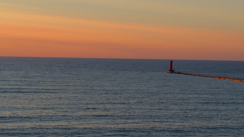 Drone aerial with a slow drift over Lake Michigan at sunrise toward the lighthouse in Sheboygan, Wisconsin, with gentle waves and golden light on the horizon.