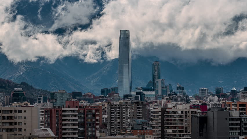 Hyperlapse with pan right movement showing the Costanera Center tower surrounded by city buildings and dramatic clouds over the Andes mountains in Santiago de Chile.
