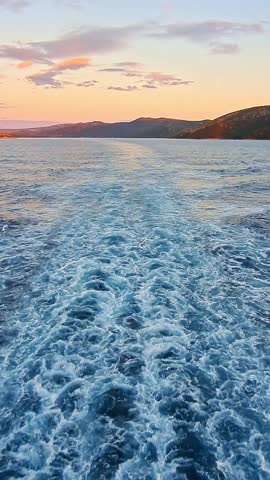 Vertical video: a serene view from the stern of a ship sailing away from the coast at sunset. The vessel sways slightly, creating a meditative mood. Ideal for social media.