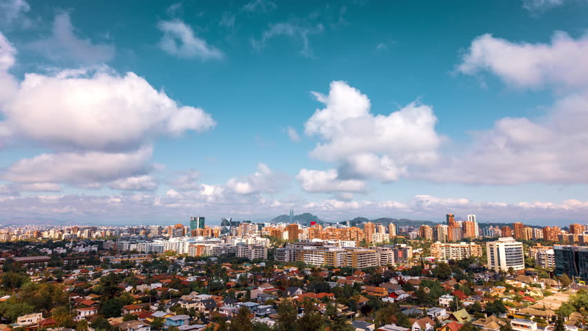 Aerial zoom in hyperlapse of Santiago city skyline with modern buildings and clouds drifting across a bright sky.