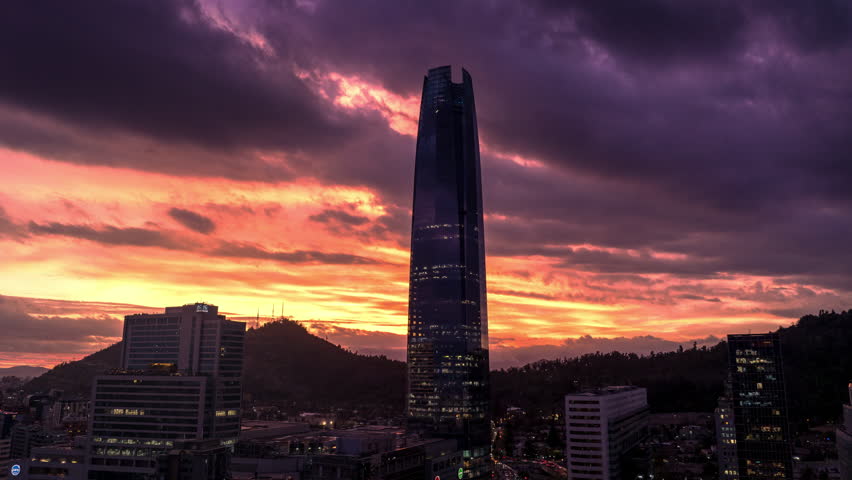 Semi orbit hyperlapse of Costanera Center surrounded by dramatic clouds and glowing sky at sunset in Santiago, Chile.