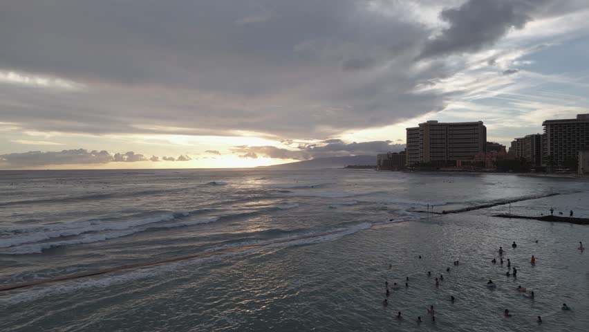 A scenic sunset view over Waikiki Beach in Honolulu, Hawaii. The ocean waves roll gently toward the shore, with surfers visible in the distance and the city skyline under dramatic evening clouds.