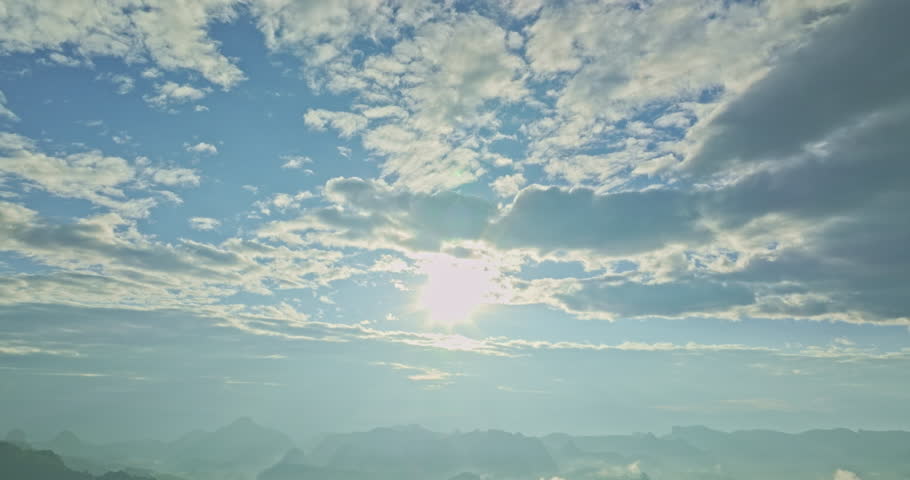  Aerial view of the sun above the mountain peaks shines through the valley. Cloudy skies and surrounding hills enhance the peaceful ambiance of this scenic setting.