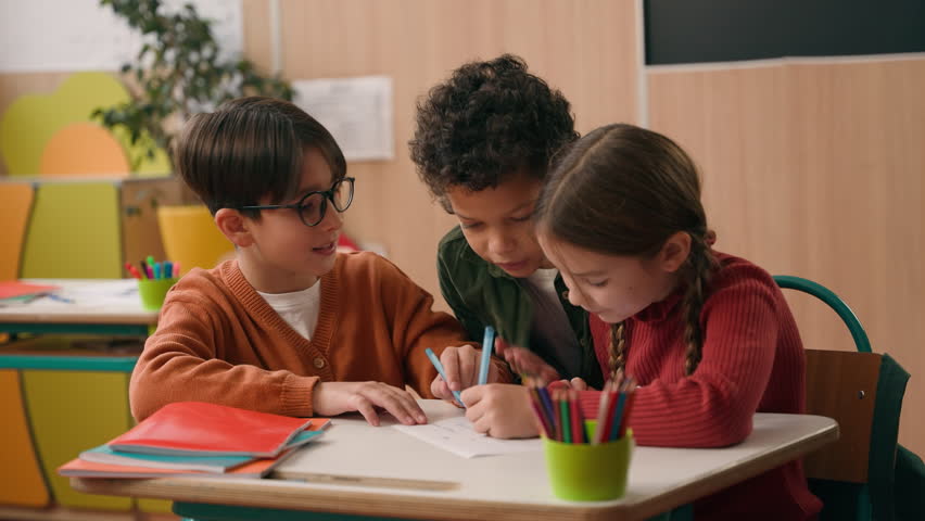 Three diversity children in school classroom two Caucasian classmates boy with girl writing drawing together African American kid child walking look at picture asking talking go away study education