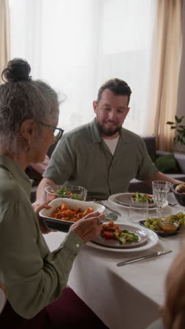 Vertical view of senior woman filling plate with sweet potatoes and passing ceramic dish to son-in-law during festive lunch at home