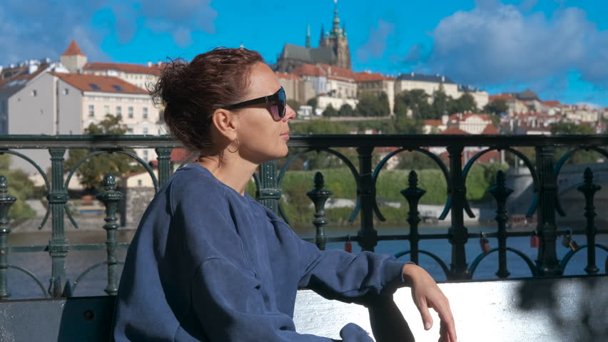 Tourist enjoying prague castle view from vltava river bank. Young woman wearing sunglasses sitting on bench overlooking historic prague castle and scenic vltava river, enjoying urban landscape