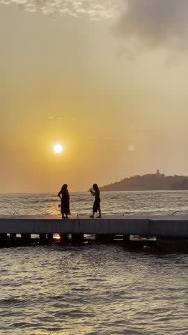 Female photographer taking photos of lady in black on pier against waving sea and sunset sky in summer evening in Kusadasi, Audin, Turkey