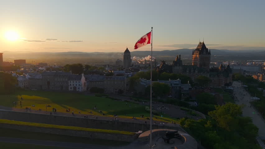 Aerial view of Quebec City at sunset featuring the Canadian flag waving above historic architecture, with warm golden light over the skyline and St Lawrence River, capturing a scenic urban landscape ideal for travel, culture, and national identity concepts