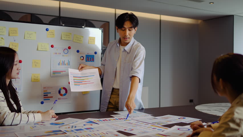 Young businessman conference speaker passionately explains business project strategy using a whiteboard, engaging a diverse team in collaborative brainstorming within a contemporary office environment