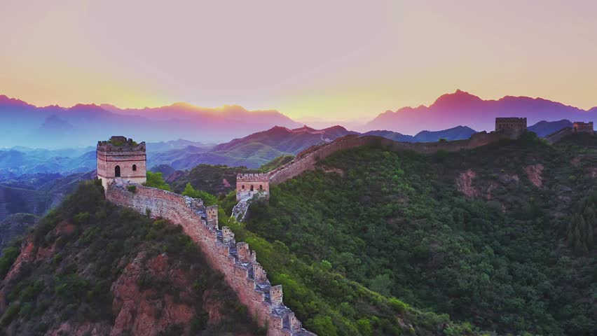A breathtaking aerial shot tracking along the Great Wall of China at sunrise, revealing ancient watchtowers atop lush mountains under a vibrant, colorful sky.