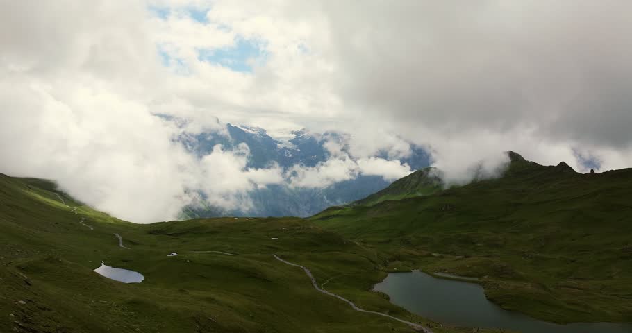Aerial shot of a dramatic alpine landscape with green valleys, mountain lakes, and winding hiking trails. Low-hanging clouds drift across the peaks, partially revealing a glacier in the distance