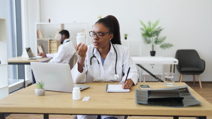 Young African medical professional in office holding pill container and writing prescription while seated at desk. Office setting shows professional environment with medical tools - Powered by Shutterstock - Get 15% off with code: PIKWIZARD15