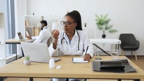 Young African medical professional in office holding pill container and writing prescription while seated at desk. Office setting shows professional environment with medical tools - Powered by Shutterstock - Get 15% off with code: PIKWIZARD15