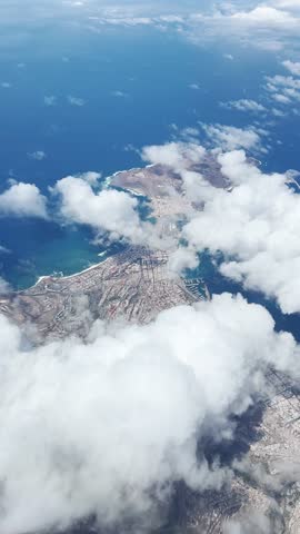 View from an airplane flying above a coastal city partially covered by clouds. Beautiful mix of urban landscape, ocean and sky from high altitude.