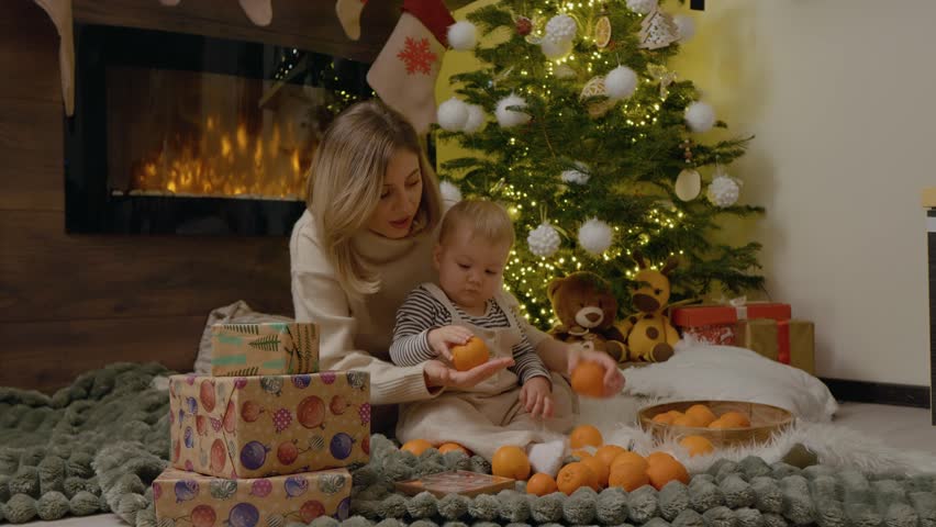 A mother and her young child sit on a cozy blanket, playing with bright oranges next to a beautifully decorated Christmas tree, creating joyful holiday memories.