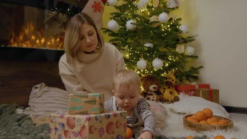 A mother and her young child share a delightful moment playing with oranges on a blanket next to a beautifully decorated Christmas tree, surrounded by festive gifts.