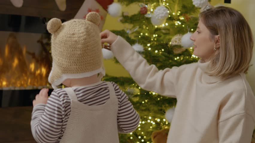 In a cozy living room, a mother helps her child put on a festive hat while they prepare to decorate the Christmas tree together with cheerful ornaments and lights.