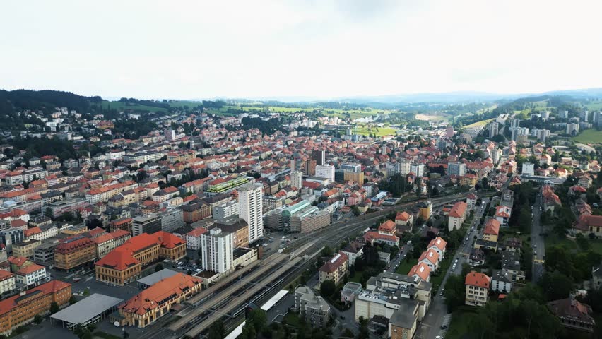Aerial view of La Chaux-de-Fonds unveils a tapestry of red rooftops, modern buildings, and railway lines amidst the lush green hills, La Chaux-de-Fonds, Neuchâtel, Switzerland.