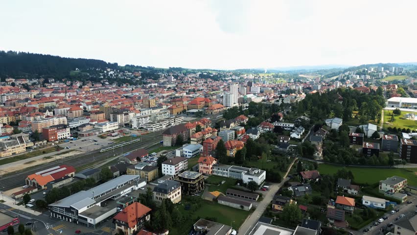 Aerial view of the cityscape reveals buildings with terracotta roofs and rail tracks cutting through, surrounded by lush trees, La Chaux-de-Fonds, Neuchâtel, Switzerland.