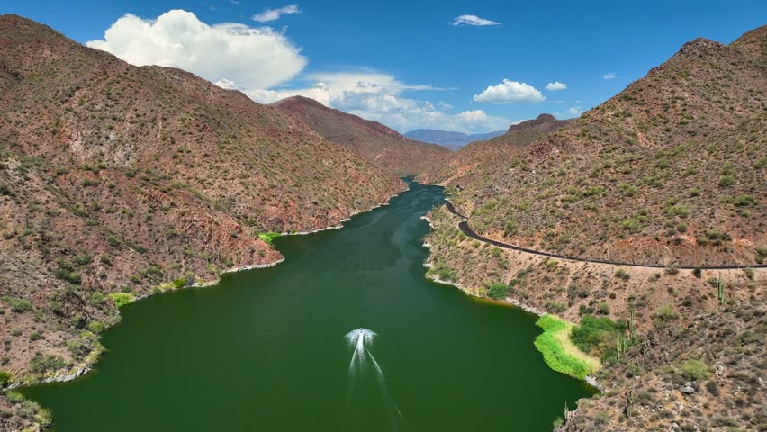 Aerial view of Theodore Roosevelt lake, where a boat glides through the green waters surrounded by the brown mountains and a winding road, Roosevelt, Arizona, United States.