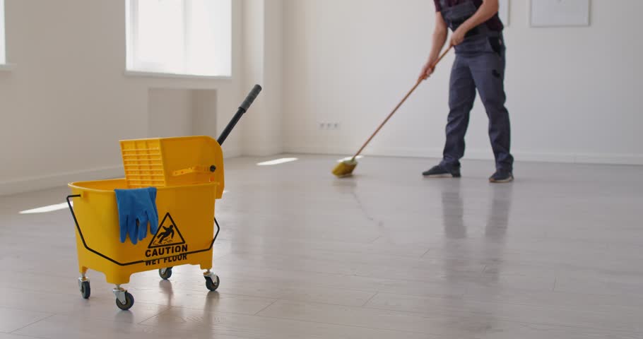 Office cleanup, workplace with janitor cleaning empty floor with broom, yellow cart with janitorial tools, caution wet floor sign. Cleaner worker in uniform holding brush to sweep dust, clean floor