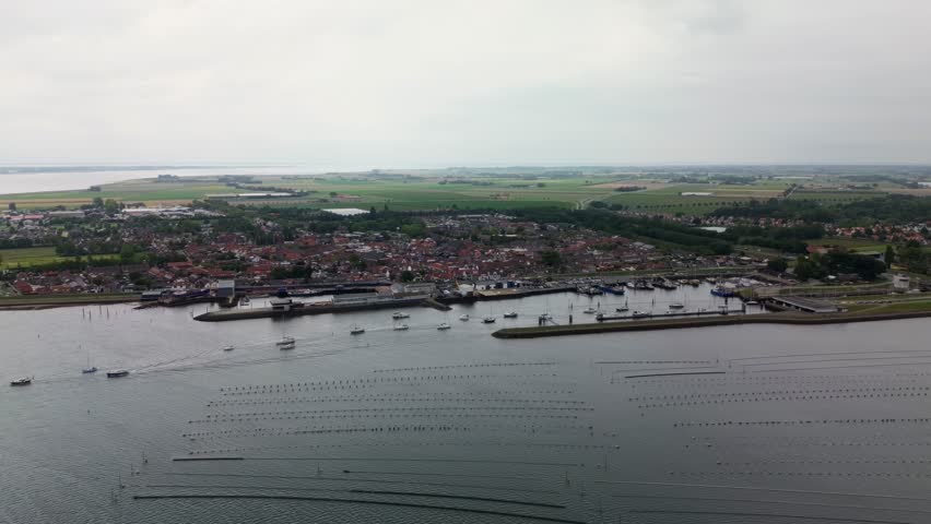 Aerial view of a coastal town with rows of buoys in the water. Boats are anchored near the harbor, and houses stretch along the shoreline under a cloudy sky.