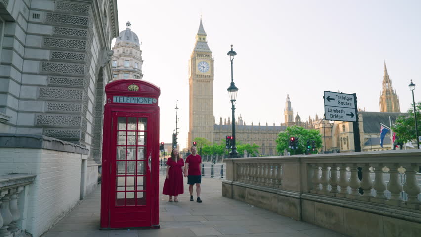 Couple Holding Hands While Walking Next To Red Telephone Box With Big Ben In The Background In London, UK. - wide shot