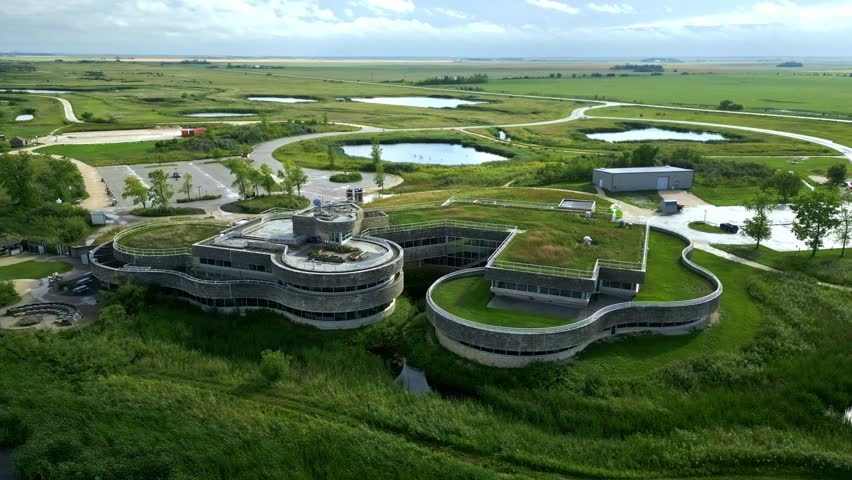 Fast-motion aerial circles modern green-roof wetland centre on a summer day