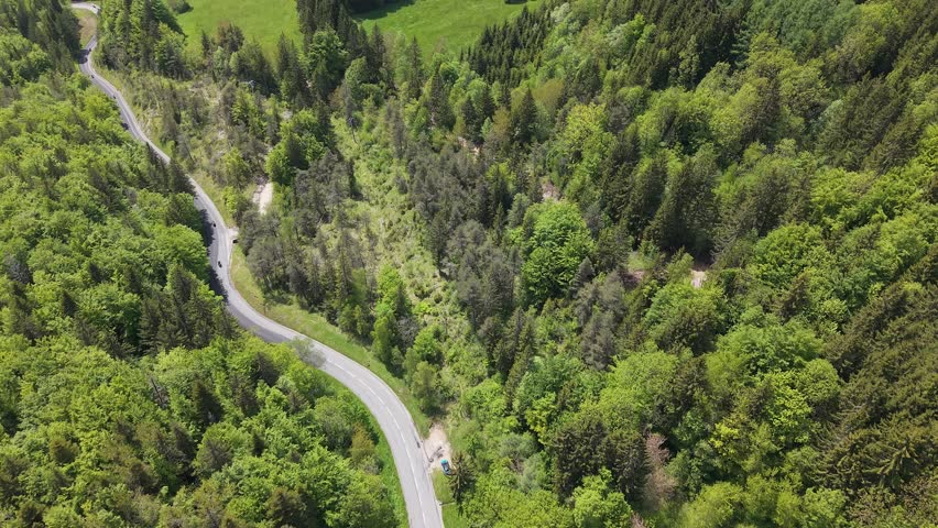 Forward drone tracking shot above road with rising gimbal, forest and mountain backdrop, French Alps, France. Scenic cinematic aerial view with cloudy dramatic sky and natural alpine landscape.